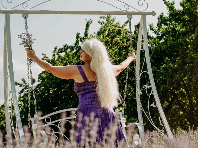 KamaliaSim, an older woman with long white hair, wearing a purple dress, holds a bouquet of lavender while standing on a white metal swing in a sunny field.