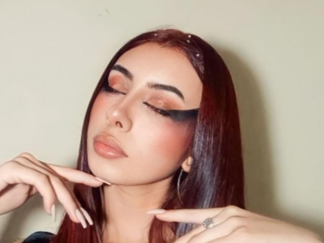 Dalila Russo ha lunghi capelli rossi, un trucco per gli occhi nero intenso e labbra rosa lucide. Le sue mani sono vicine al viso e ha un piccolo tatuaggio sul dito indice destro..