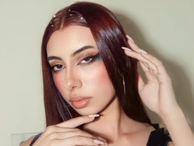 Dalila Russo, mit ihren langen roten Haaren, dem auffälligen Make-up und den glänzenden Lippen, berührt ihr Kinn mit ihren gepflegten Fingern und hat einen verführerischen, sinnlichen Ausdruck. Ihre Brüste sind teilweise sichtbar..