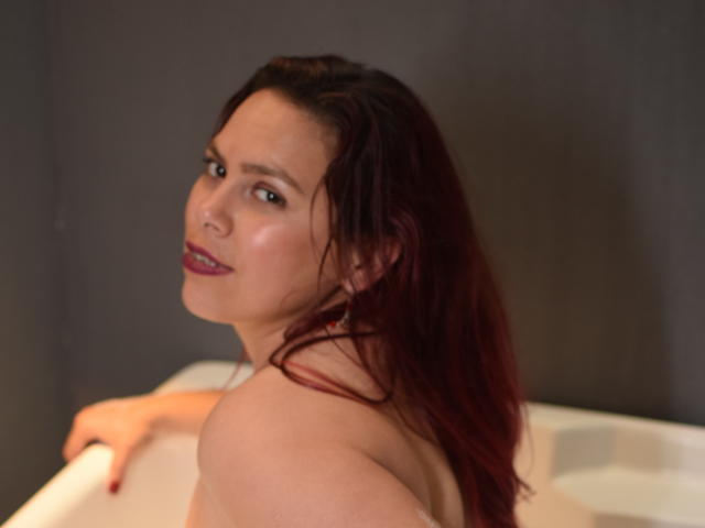 Salma Brown, com cabelo ruivo, olhando por cima do ombro, sem blusa, em uma banheira, mostrando suas costas nuas e seios pequenos e firmes. Ela está sorrindo levemente, com batom vermelho..