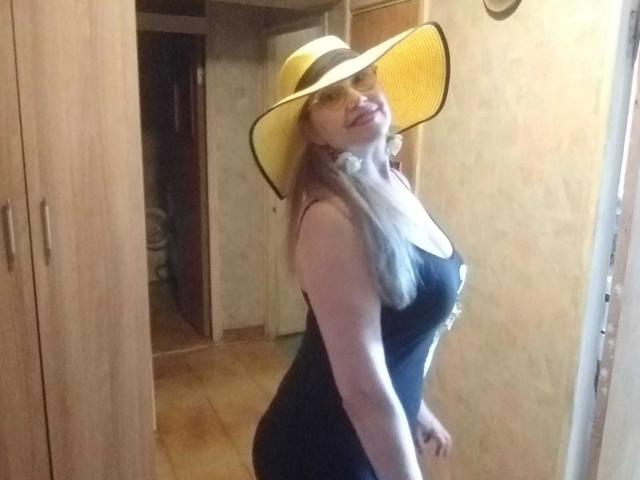 LadyMariahX with big tits, wearing a black dress and yellow sun hat, standing in a room, smiling, blonde hair, fair skin, and curvy body, with her ass slightly turned.