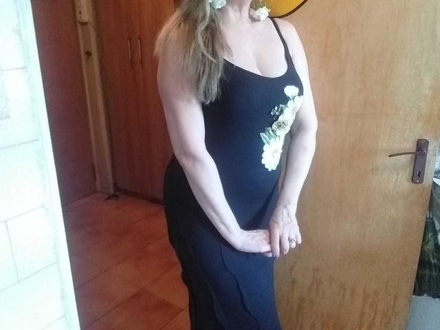 LadyMariahX in a black tank top and black skirt, with white floral design, standing in a wooden room, hands near her crotch, visible breasts, blonde hair in a ponytail.