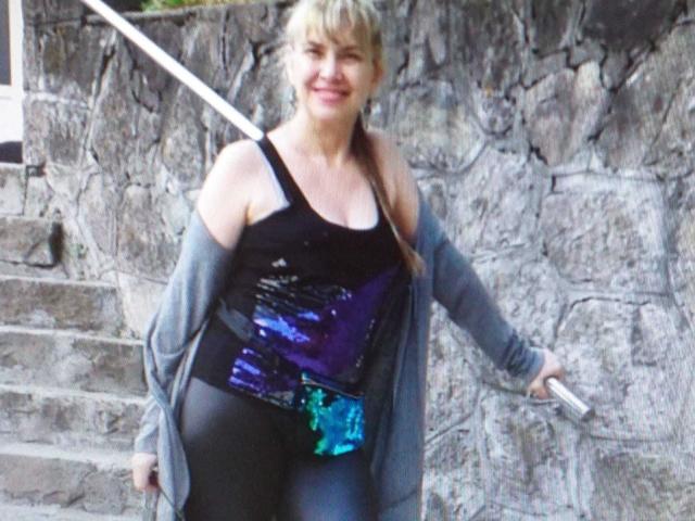 LadyMariahX with blonde hair, smiling, wearing a black tank top with purple and blue sequins, gray cardigan, and black pants, holding a white cane, standing on stone steps.