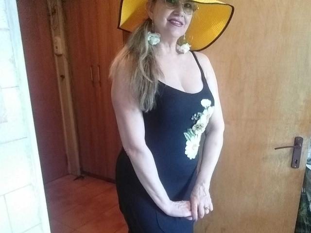 LadyMariahX with pale skin, blonde hair, wearing a yellow hat, black tank top showing her tits, and black pants, smiling, hands together, wood door in background.