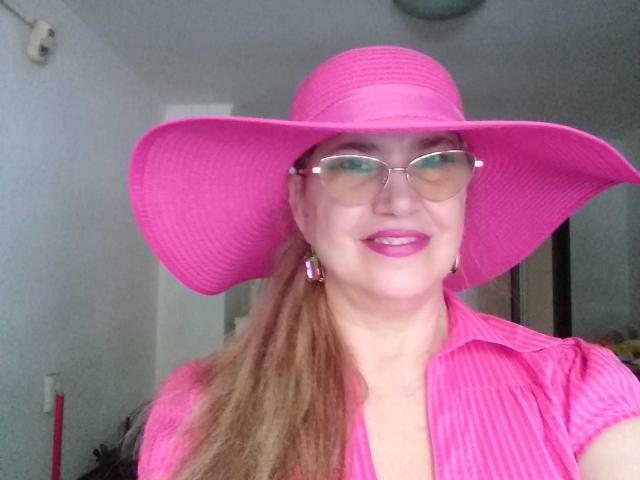 LadyMariahX with pink hat, glasses, and pink shirt, showing off her tits, pink lipstick, and long brown ponytail, smiling in a selfie.