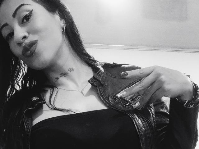 Autoportrait en noir et blanc de LunaCandy, avec des cheveux noirs et mouillés, des lèvres pincées, un anneau au nez et un piercing aux lèvres, portant une veste en cuir sur les seins, une main sur la poitrine, et un tatouage au cou..