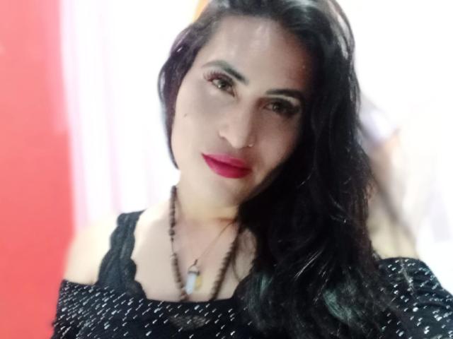 ElectraHottyX ha i capelli scuri, il rossetto rosso e indossa una maglietta nera con una spalla scoperta, con una collana di perline, che mette in mostra le sue spalle scoperte e un piccolo anello al naso..