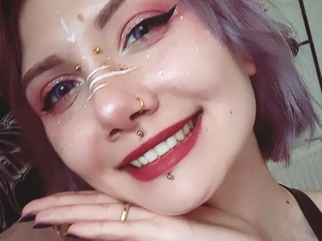 PerfectGabbie, con cabello morado, lápiz labial rojo y múltiples perforaciones faciales, sonriendo con un líquido blanco en su rostro, con la mano apoyada en la barbilla, vistiendo una blusa negra..