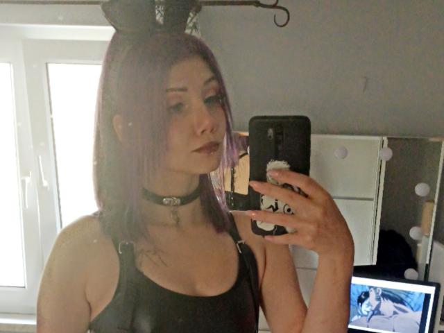 PerfectGabbie con cabello morado, camiseta negra, collar, sosteniendo un teléfono, selfie frente al espejo, pechos pequeños, piel pálida, maquillaje oscuro, una laptop en el fondo mostrando a una mujer de cabello azul..