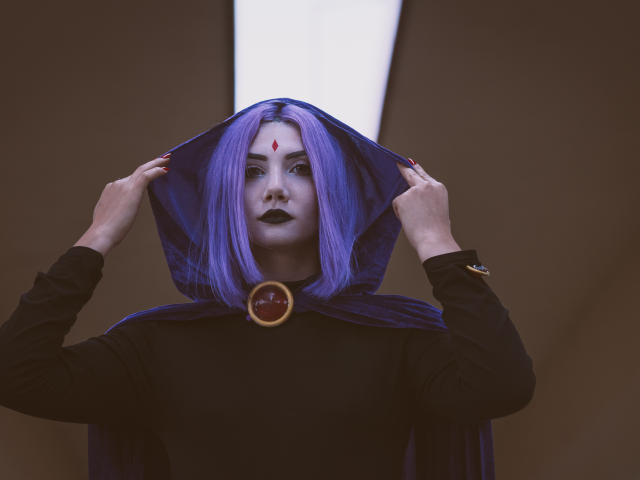 PerfectGabbie with purple hair, black lipstick, and a red gem on her forehead, holding up a blue hooded cloak, wearing a black top, looking intense.
