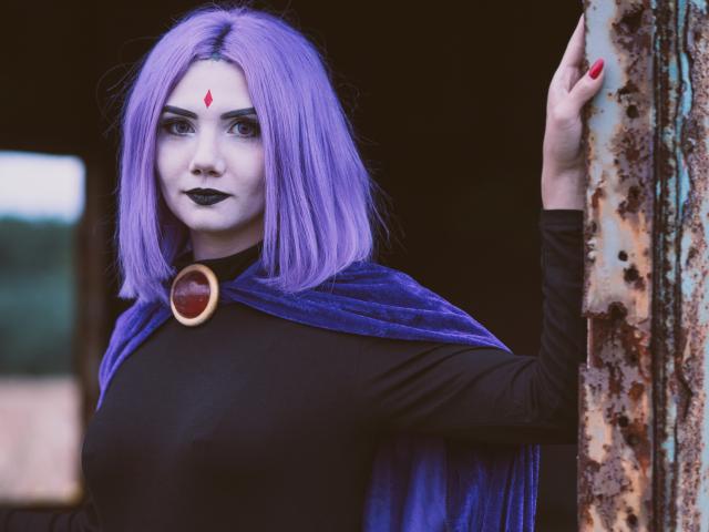 PerfectGabbie with purple hair, black makeup, red gem on forehead, blue cape, black top, holding rusty door. Boobs visible under top, red nail polish, serious expression.