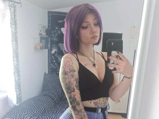 PerfectGabbie con un corte de pelo morado, top corto negro, mostrando tatuajes en los brazos y el torso, sosteniendo un teléfono, mirando hacia abajo, usando una falda de cuadros, de pie frente a un espejo..
