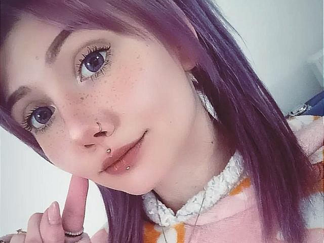 PerfectGabbie has purple hair, blue eyes, freckles, a nose ring, lip piercing, and is pointing at her face with a finger. She's wearing a pink, fuzzy, patterned top.