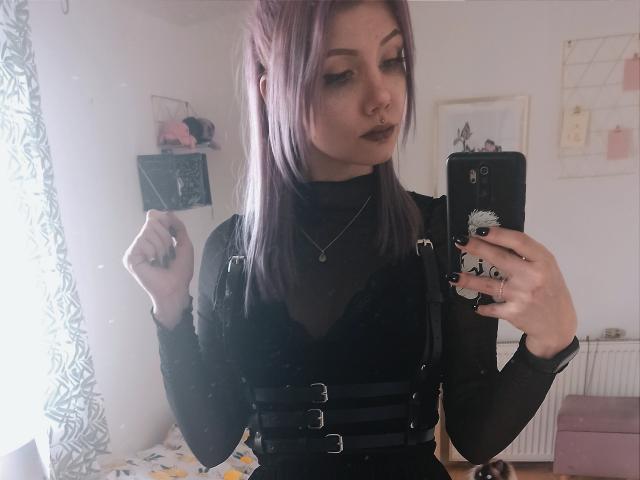 PerfectGabbie met paars haar, zwarte lipstick en een zwarte jurk, die een telefoon vasthoudt, zwarte nagels laat zien en een neusring draagt, in een spiegelzelfportret. De borsten zijn bedekt door de bandjes van de jurk..