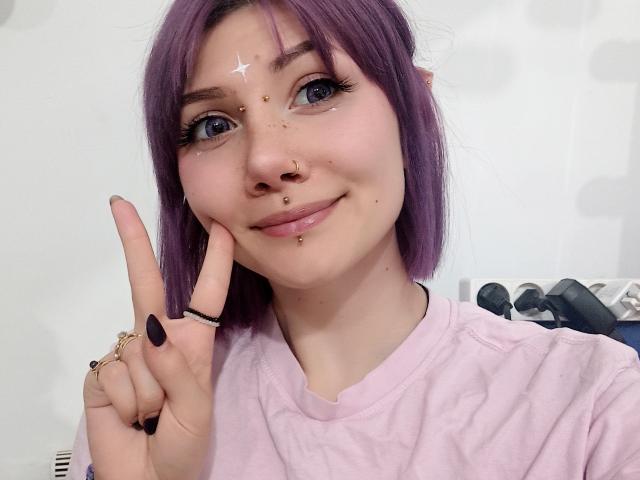 PerfectGabbie, con cabello morado, ojos azules y múltiples perforaciones faciales, sonriendo, haciendo un gesto de la paz, vistiendo una camisa rosa y con una apariencia linda y un poco nerd..