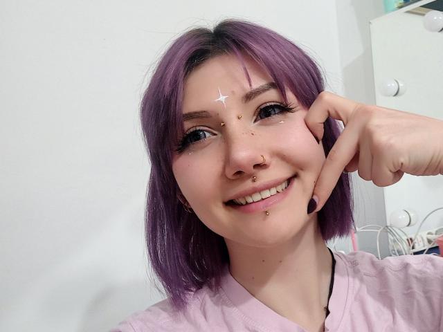 PerfectGabbie, con cabello morado, sonriendo, tiene maquillaje con estrellas y puntos en la frente, un piercing en la nariz, un piercing en el tabique nasal, un piercing labial y está tocándose la mejilla. Lleva una camisa rosa, con un fondo de pared blanca..