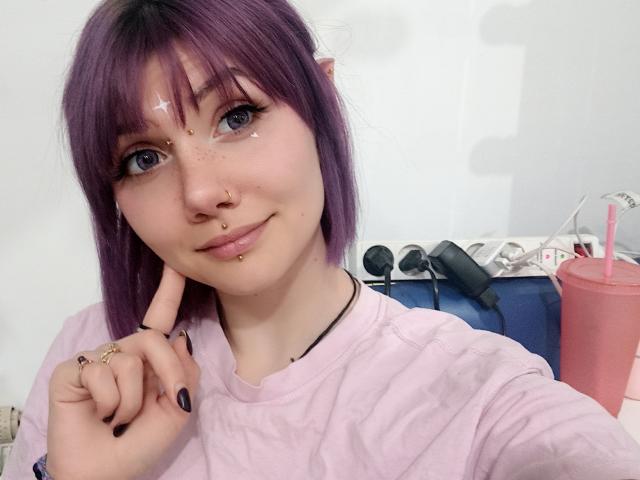 PerfectGabbie with purple hair, nose ring, and septum piercing, touching her chin with a finger, wearing a pink shirt, freckles, and black nail polish, smiling softly.