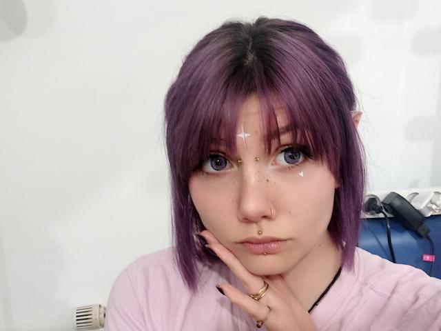 PerfectGabbie has purple hair, blue eyes, and a nose ring. She's wearing a pink shirt, with a hand resting on her chin, and has sparkles on her face.