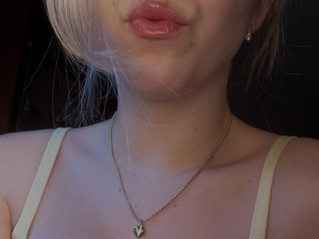 SugarrXs rosige, glänzende Lippen, helle Haut, blondes Haar, eine kleine Halskette mit einem Schmetterlingsanhänger und dünne Spaghetti-Träger an einem beige farbenen Oberteil. Die Brüste sind teilweise sichtbar..