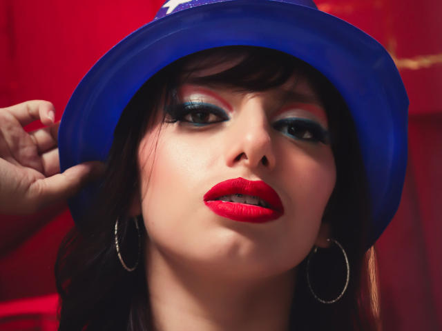 LeticiaFlorenz has bold red lipstick, blue hat, and green eye makeup, with hoop earrings, looking seductive, her hand holding the hat, red background.