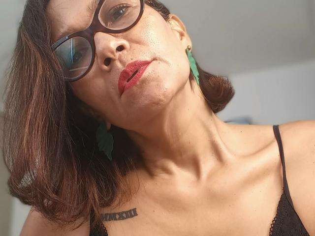 JennyX, de aspecto maduro, con labios rojos, gafas negras, pendientes verdes y una blusa negra de encaje, mostrando una expresión ligeramente fruncida, con sus pechos y clavícula visibles. Su cabello castaño le llega a los hombros..