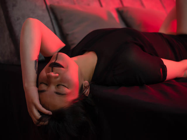 SofiaHazard acostada en una cama, vestido negro, mano en la cabeza, ojos cerrados, iluminación roja y rosa, lápiz labial negro, relajada, sensual, suave, sábanas de la cama, oscura, melancólica..
