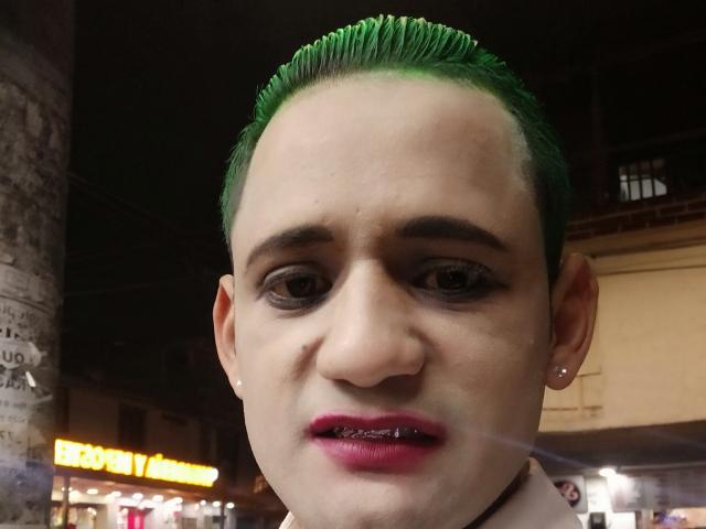 KimJimmy has green-slicked-up hair, dark eyebrows, pink lipstick, and a piercing in each ear. Their face is slightly squished with a small, visible tooth. Nighttime.