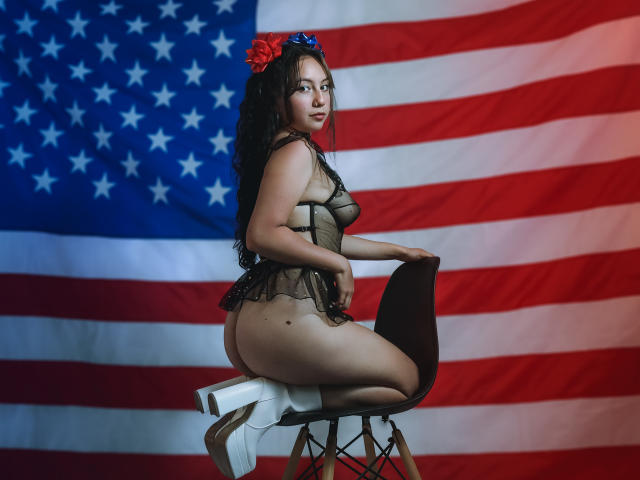 VeroHotLove, formosa con un grande sedere, indossa lingerie nera trasparente e tacchi bianchi, seduta su una sedia, capelli neri, fiore rosso e blu, sfondo con la bandiera americana, sexy, sicura di sé, che guarda indietro..