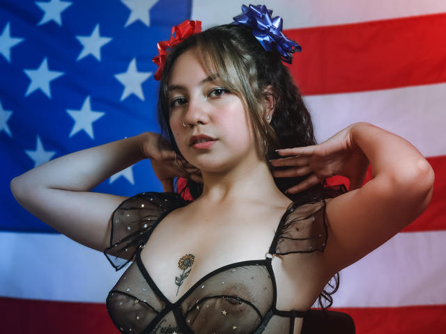 VeroHotLove, con cabello largo y castaño, vistiendo una blusa negra transparente que muestra sus senos, lazos para el cabello azules y rojos, un piercing en la nariz y un tatuaje en el pecho, con las manos detrás de la cabeza..