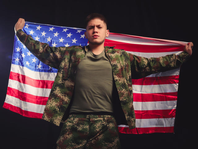 SebastiianMaxime está de braços abertos, segurando uma bandeira americana, vestindo uma jaqueta e calças de camuflagem verde, com uma expressão séria, cabelo castanho curto, fundo preto..