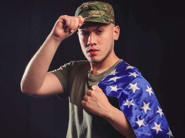 SebastiianMaxime, grüne Tarnmütze, grünes T-Shirt, hält eine blaue amerikanische Flagge mit weißen Sternen, hebt den Faust zum Kopf, ernster Gesichtsausdruck, dunkler Hintergrund, kurzer Haarschnitt, kleines Ohrpiercing..
