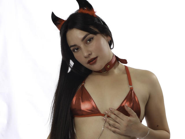 Julianal met lang zwart haar, rode duivelhoorns en een glanzend rood bikini topje, waarbij ze haar kleine borsten met één hand bedekt, draagt een rode choker en een armband..