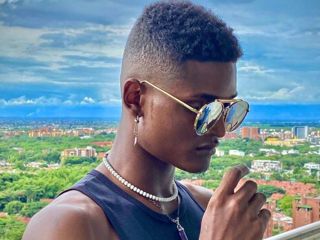 James Makony, con i capelli neri ricci e corti, indossa occhiali da sole aviator dorati, una canottiera nera, una collana di perle e un orecchino, tiene in mano una sigaretta, sullo sfondo di un cielo blu con la città e la vegetazione sottostanti..