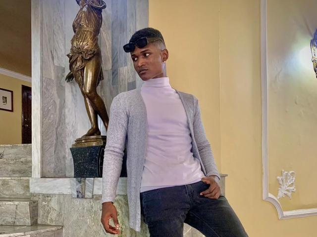 JamesMakony está em pé, com uma expressão séria, vestindo um cardigã cinza, uma gola alta branca e calças jeans pretas, apoiado em uma estátua. Ele está usando óculos de sol na cabeça e tem cabelo curto..