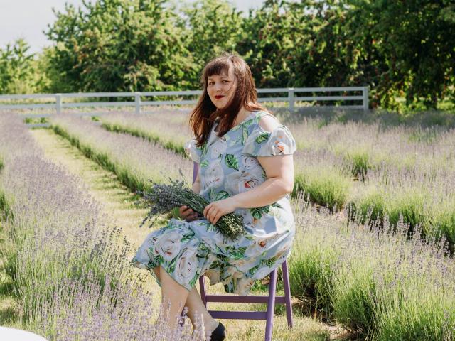 AliceInks with brown hair, wearing a floral dress, sitting on a purple chair in a lavender field, holding a bouquet of lavender, smiling with red lipstick.