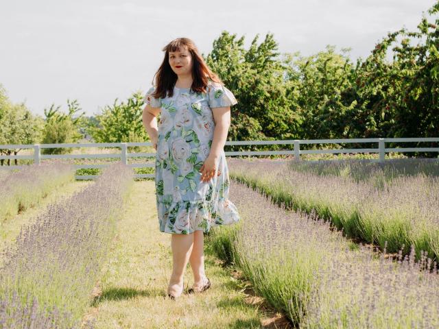 AliceInks stands in a lavender field, wearing a floral dress, holding her hand near her ass, with green trees and a white fence in the background.