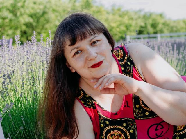 AliceInks with long brown hair, red lipstick, freckles, and a pink dress with black and gold patterns, resting her chin on her hand, smiling in a lavender field.