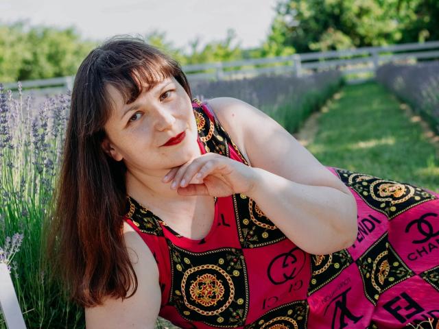 AliceInks, a chubby woman with pale skin, brown hair, and red lipstick, wears a red and black patterned dress, leaning on her hand, in a lavender garden.