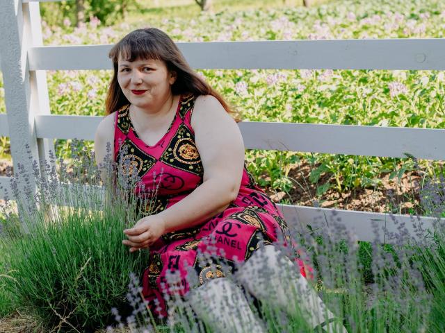 AliceInks, chubby with brown hair, sitting on white fence, wearing pink and black patterned dress, smiling, surrounded by green grass and purple flowers, looking relaxed and happy.