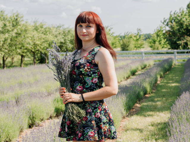 NinaFlawlees with red hair, holding a bouquet of lavender, wearing a floral dress, standing in a lavender field, looking slightly to the side, under a sunny sky.