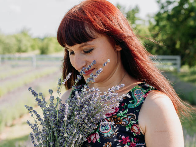 NinaFlawlees with red hair, holding a bunch of lavender, wearing a colorful floral dress, looking down with a gentle smile, soft sunlight on her face, outdoor setting.