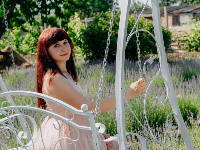 NinaFlawlees with long red hair, pink dress, sitting on white metal swing, holding chain, looking up, in sunlit lavender field. Her tits are subtle, her ass is unseen.