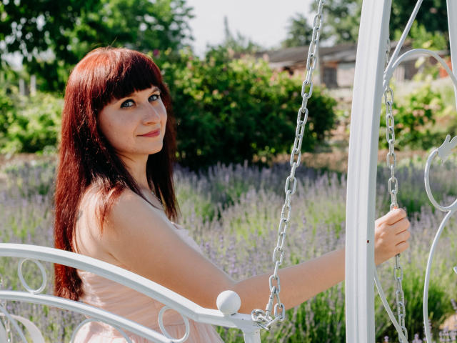 NinaFlawlees with red hair, holding white metal swing chains, wearing a pink dress, looking back with a slight smile, standing in a lavender field.
