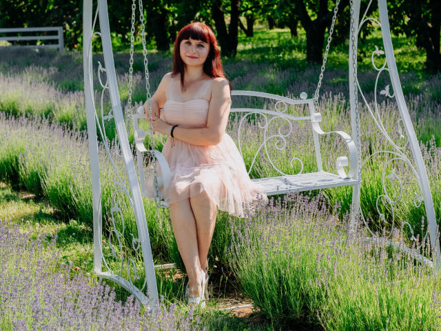 NinaFlawlees, with red hair, sits on a white, ornate swing in a lavender field, wearing a pink, strapless, tulle dress, arms crossed, smiling, legs together, in sunlight.