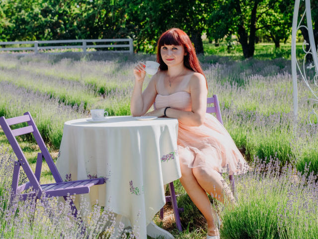 NinaFlawlees with red hair, pink strapless dress, holding white cup, sitting at purple table with white cloth, green grass, and lavender, sunny outdoor setting. Her ass is visible.