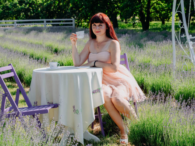 NinaFlawlees, com cabelo ruivo, pele clara, seios pequenos, vestindo um vestido rosa, sentada em uma mesa roxa, segurando uma xícara de chá branca, em um campo de lavanda..