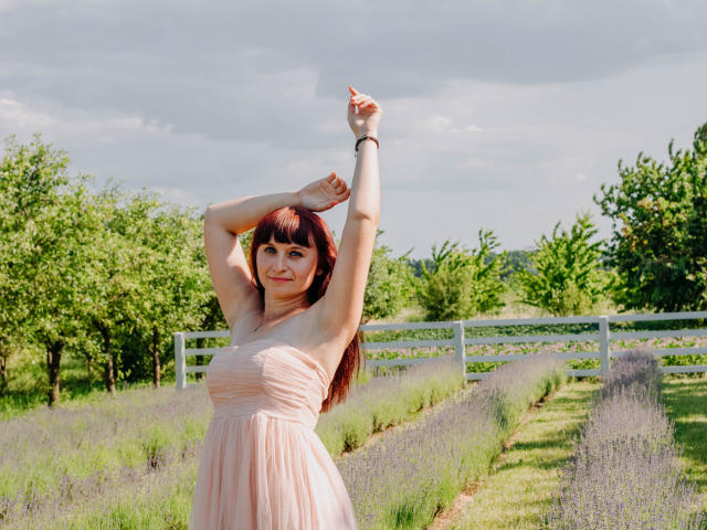 NinaFlawlees met armen omhoog, gekleed in een roze jurk, staand in een lavendelveld, lachend, met rood haar en een armband, onder een bewolkte lucht..