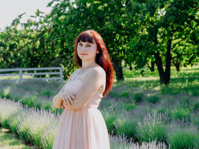 NinaFlawlees with red hair, crossed arms, wearing a pink, strapless dress, standing in a lavender field, green trees in background, sunny, smiling, freckles.