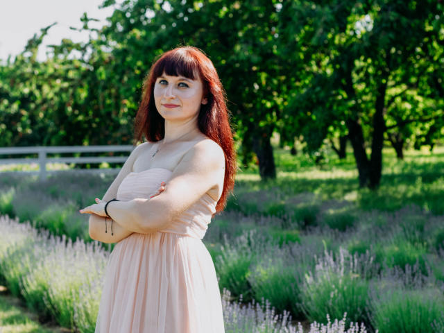 NinaFlawlees stands with arms crossed, wearing a pink dress, showing her tits, with red hair and a calm expression in a sunny lavender field.