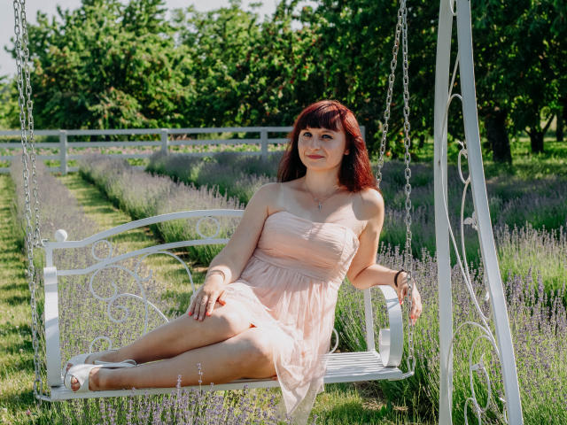 NinaFlawlees, con cabello rojo, piel pálida y senos pequeños, vestida con un vestido rosa, sentada en un columpio blanco y ornamentado, en un campo de lavanda, con árboles verdes en el fondo..